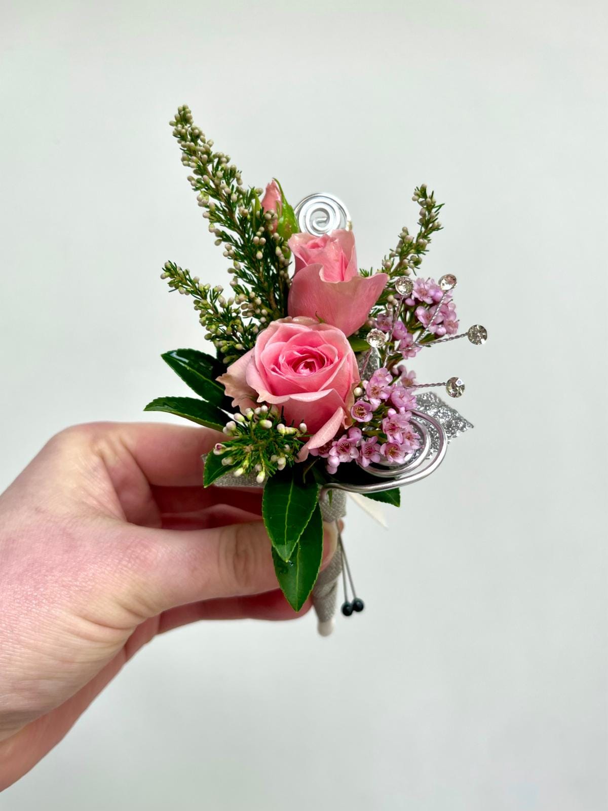 Small pink rose boutonniere with delicate filler flowers and silver accents held in a hand