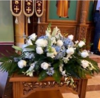 Blue and White casket spray - A serene, altar-style casket spray in white, blue, and green that brings calm dignity to a funeral or memorial service. This low, elongated arrangement of white roses and pale blue hydrangeas-built for a casket or church altar-creates a quietly reverent focal point that honors the person you're thinking of and supports a reflective ceremony.  Rounded clusters of blue hydrangeas form the center, surrounded by tightly clustered white roses and taller white snapdragons that add gentle vertical texture while preserving the horizontal profile. Broad glossy leaves and ferns frame the blooms and spill over the edges giving the piece a finished, formal presence suited to religious services and memorial gatherings.  Designed to sit low across a casket or on an altar, this composition emphasizes a peaceful, respectful mood and reads well from all sides during a service.