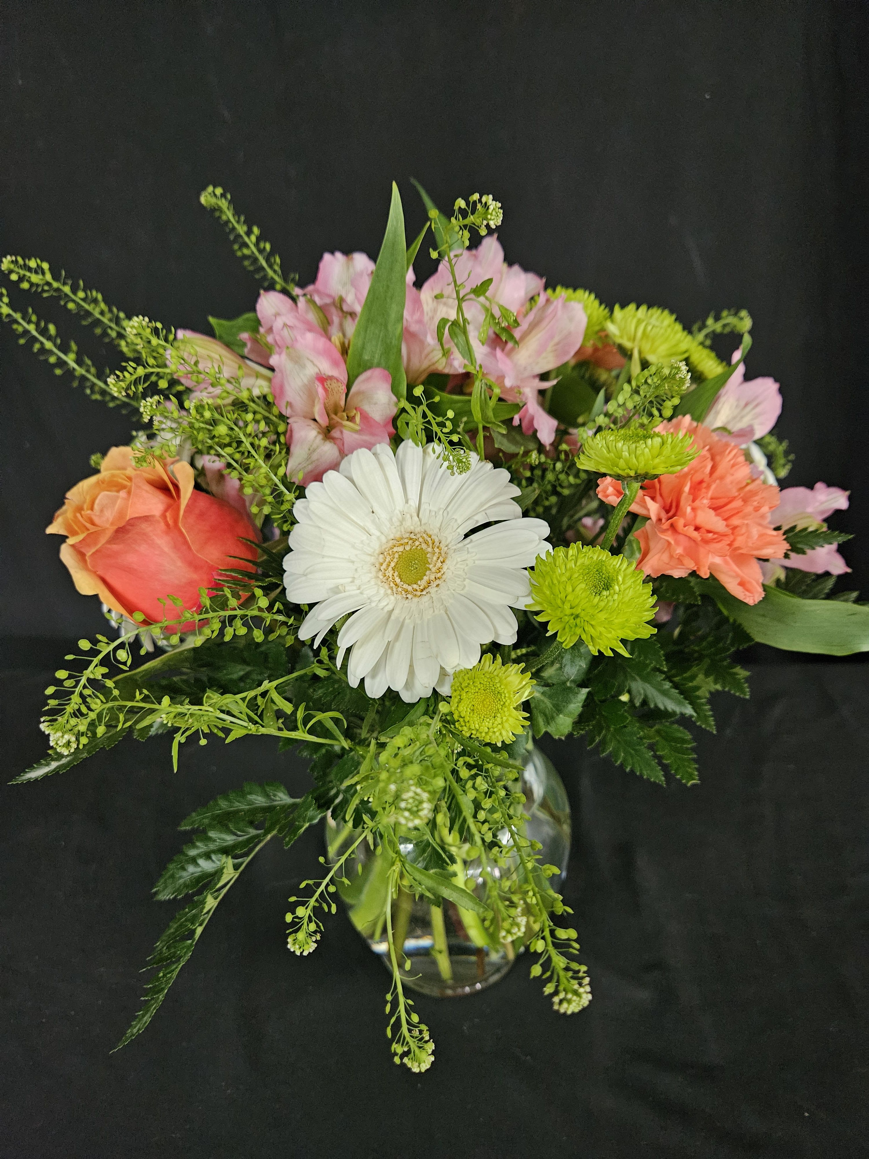 Mixed bouquet with white gerbera daisy, orange rose, pink blooms, and green mums in a clear glass vase.