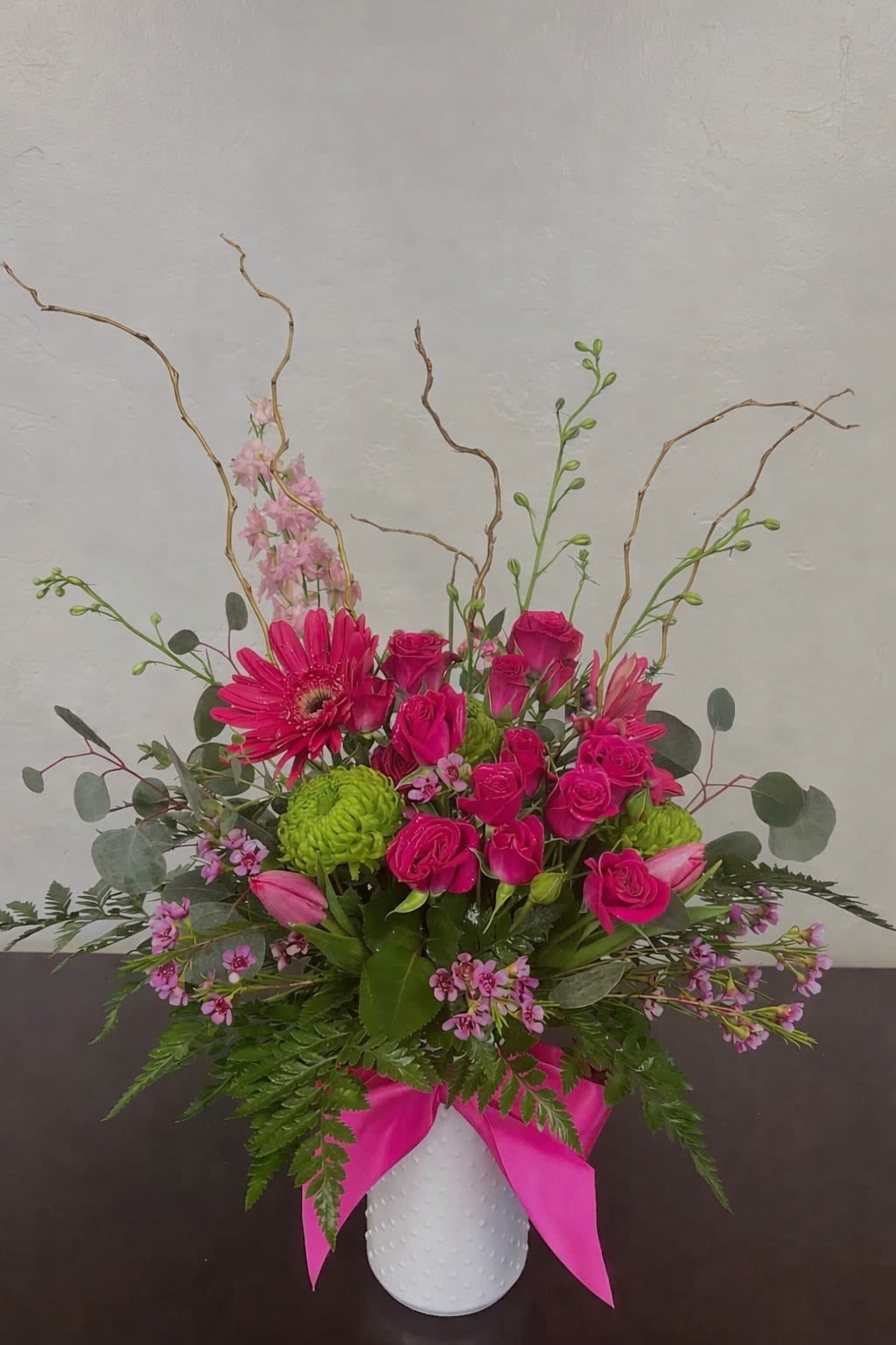 Fuchsia Garden Celebration - Vibrant, joyful, and full of personality, Fuchsia Garden Celebration is designed to make a statement. Featuring lush hot pink spray roses, bold gerbera daisies, fresh tulips, and bright green button mums, this arrangement bursts with color and life. Accented with delicate wax flower, airy greenery, and sculptural curly willow, the design brings a modern garden feel with artistic movement and texture. The crisp white ceramic vase paired with a striking fuchsia satin ribbon adds a polished, upscale finish. Perfect for birthdays, celebrations, or simply sending a little happiness, this arrangement is handcrafted with premium blooms and designed to stand out—because ordinary just isn’t your style.