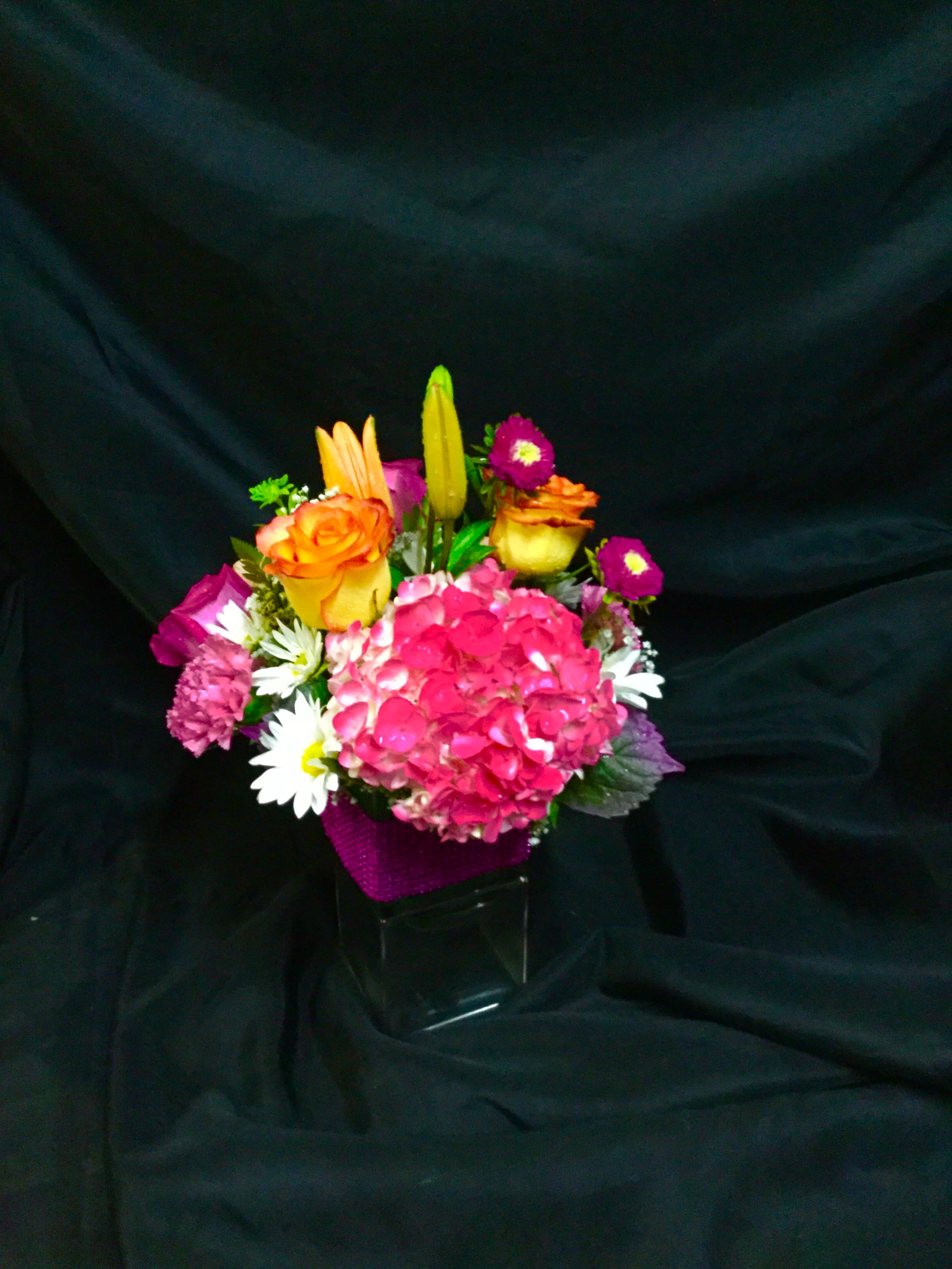 Bright mixed bouquet with pink hydrangea, orange roses, lilies, and daisies in a clear cube vase with purple wrap