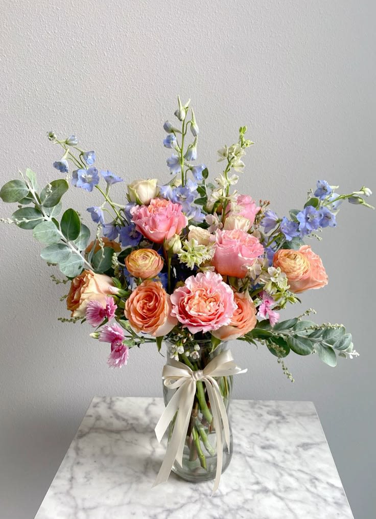Cottage Sky - Soft and romantic, Cottage Sky features lush peach and blush roses accented with delicate blue delphinium, roses, ranunculus and fresh eucalyptus in a clear glass vase finished with a satin ribbon. Its airy, garden-style design brings together gentle pastel tones for a timeless arrangement perfect for birthdays, sympathy, new babies, or simply sending a thoughtful touch.  Please note: Flower varieties and colors may vary based on seasonal availability. Substitutions may be made with flowers of equal or greater value while preserving the overall style, shape, and color palette of the arrangement.