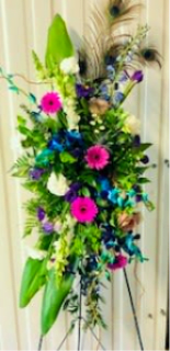 Exquisite Tribute - A tall, cascading standing spray that makes a dignified statement - Exquisite Tribute is a sympathy and funeral flower arrangement designed to convey heartfelt remembrance. Pink gerbera daisies and soft roses offer warmth and compassion while blue and teal dendrobium orchids lend a tranquil, uplifting presence; the vertical composition reads like a memorable salute at a memorial service or celebration of life.  This arrangement is mounted on a metal easel and built around glossy tropical foliage that frames clusters of snapdragons, spiked white blooms, and smaller purple and cream flowers for layered texture and depth. Wispy peacock feathers extend from the top to add dramatic height and a refined focal point, giving the piece a formal, expressive silhouette ideal for placing beside a casket or at a service entrance.  Appropriate for honoring a dearly missed loved one, this standing spray communicates respect and thoughtful care for the person you're remembering.