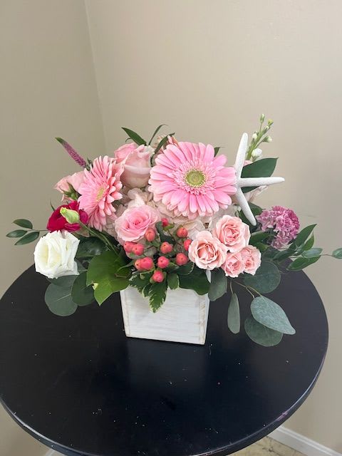 Pink gerbera daisies, roses, and spray roses in a white wooden cube with a decorative starfish