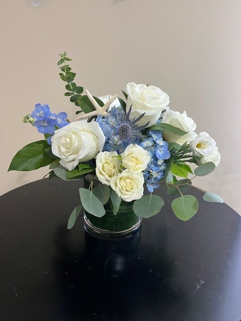 Low arrangement of white roses and blue flowers in a clear glass vase with a small starfish accent.