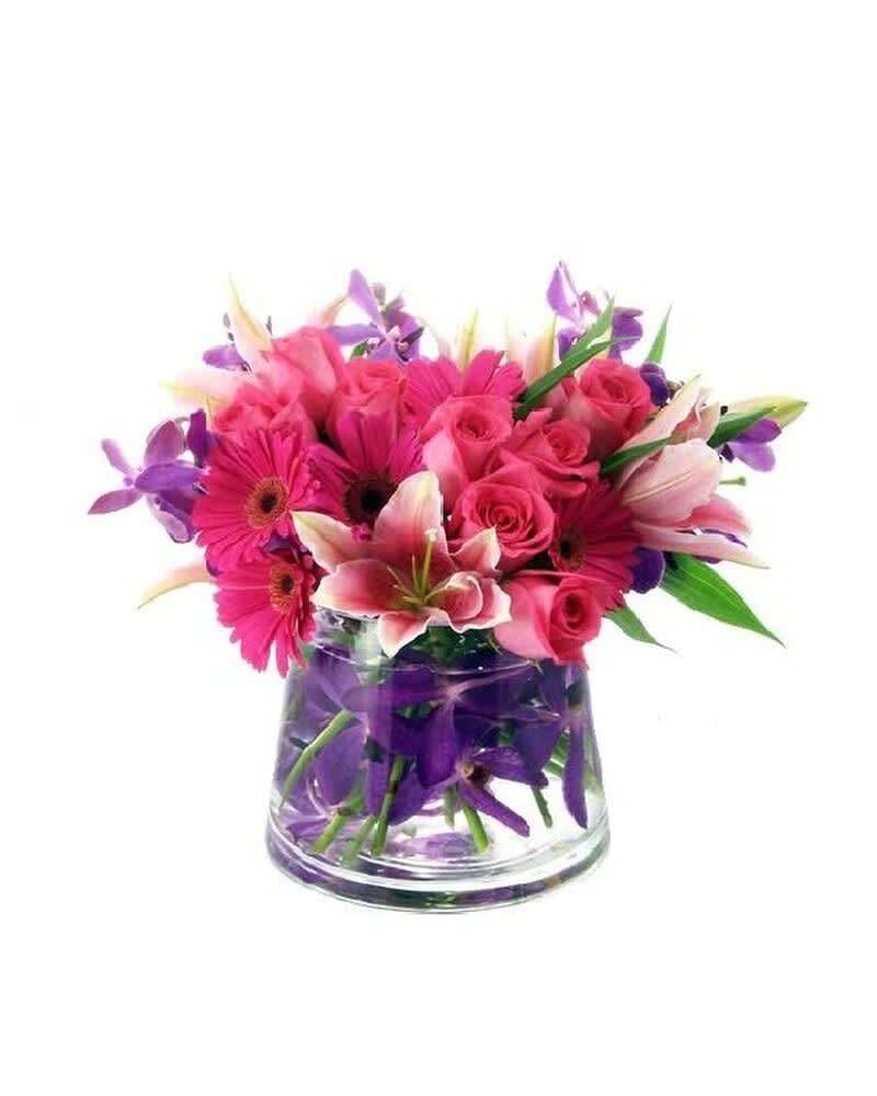 Low arrangement of pink gerbera daisies, roses, and lilies with purple orchids in a clear glass vase