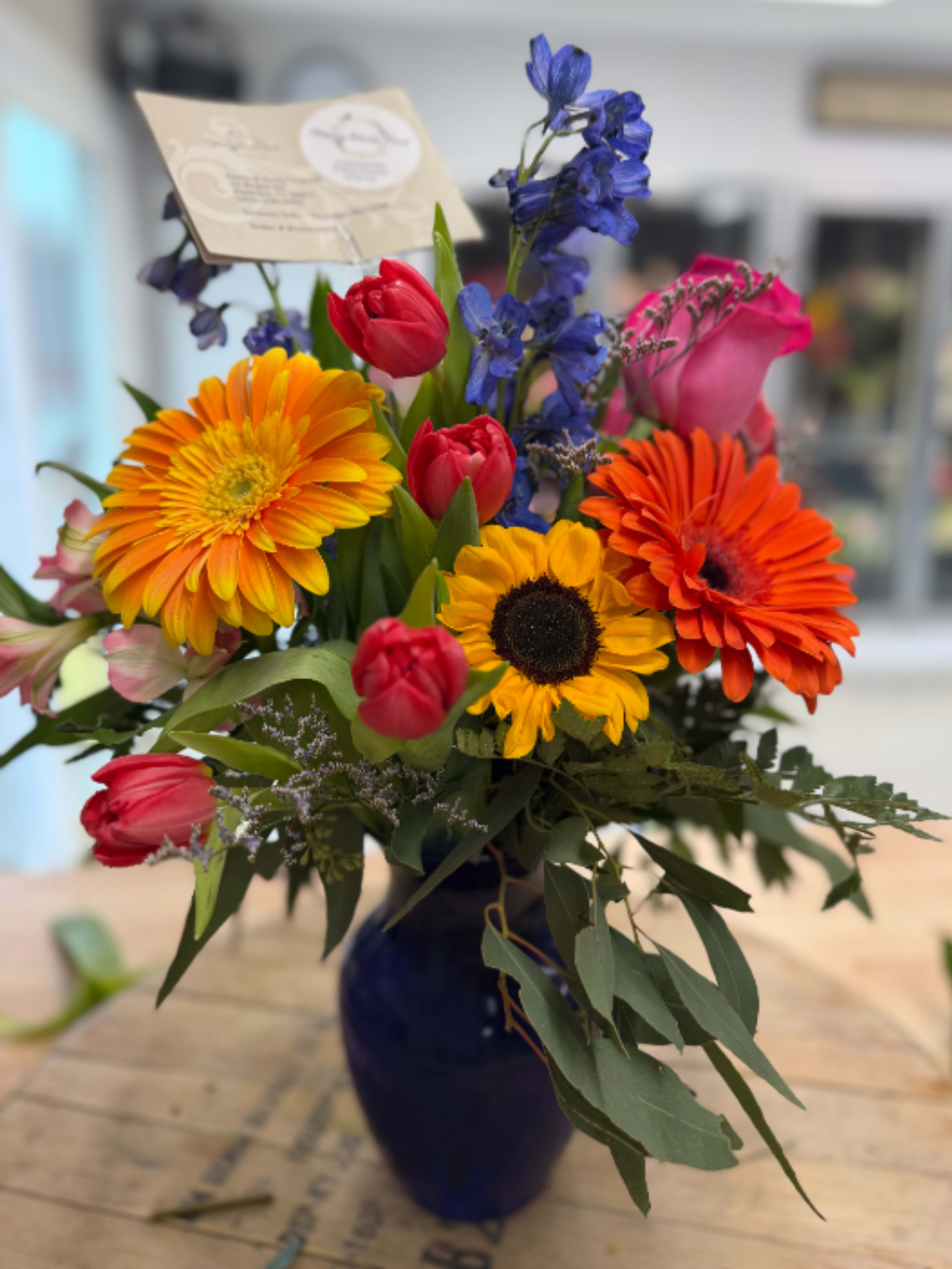 Bright mixed bouquet with orange gerbera daisies, a yellow sunflower, and pink tulips in a blue vase.