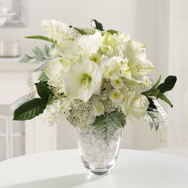 White amaryllis, roses, and small daisies in a clear glass vase with ribbon accents