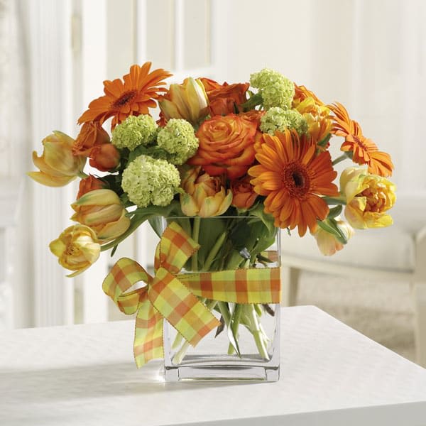 Bright orange and yellow bouquet with gerbera daisies, roses, and tulips in a square glass vase with plaid ribbon.