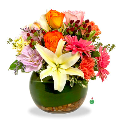 Colorful arrangement of lilies, roses, gerbera daisies, and carnations in a round green glass vase with pebbles.