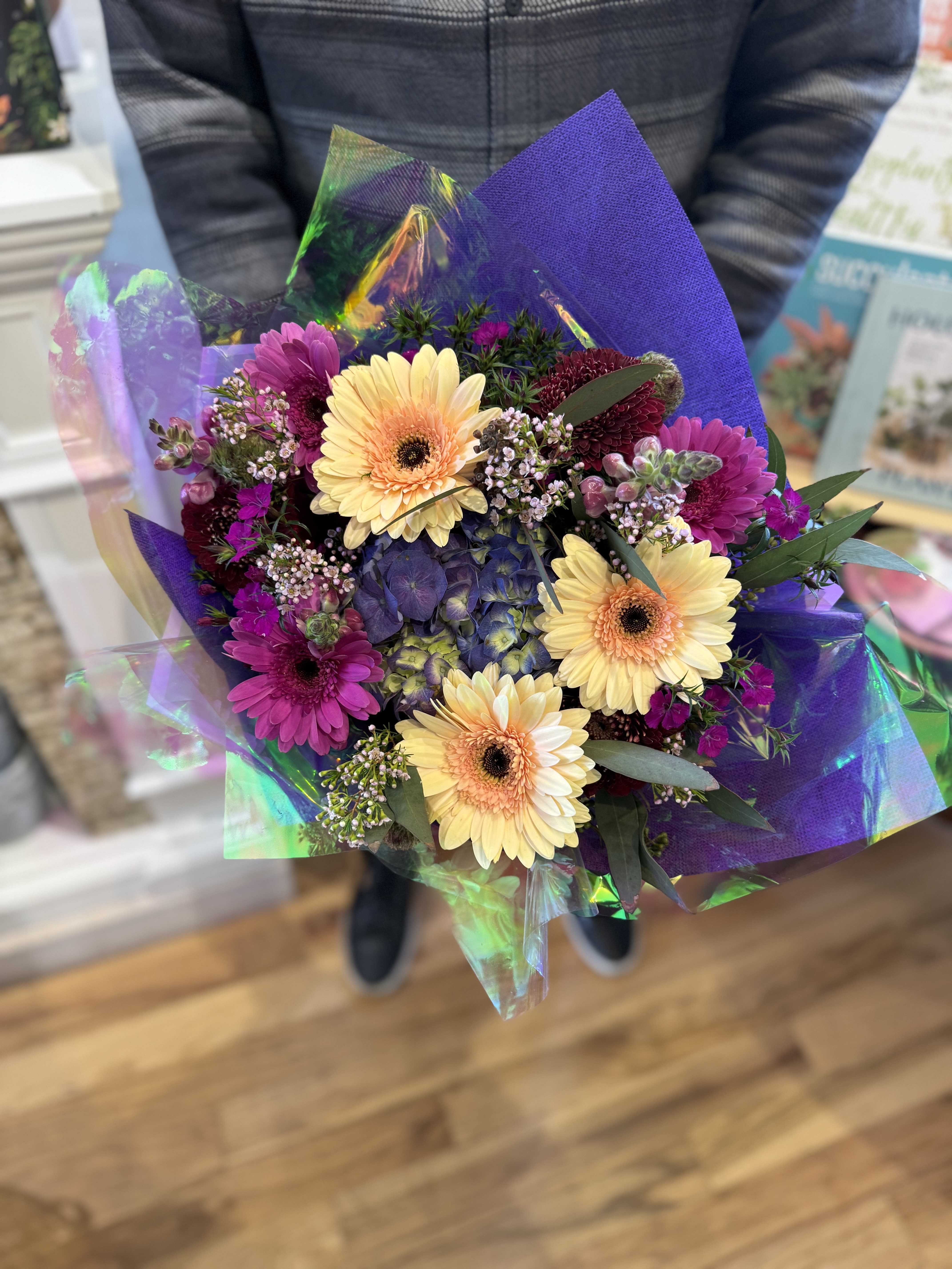 Hand-tied bouquet of yellow gerbera daisies and purple flowers wrapped in iridescent cellophane.