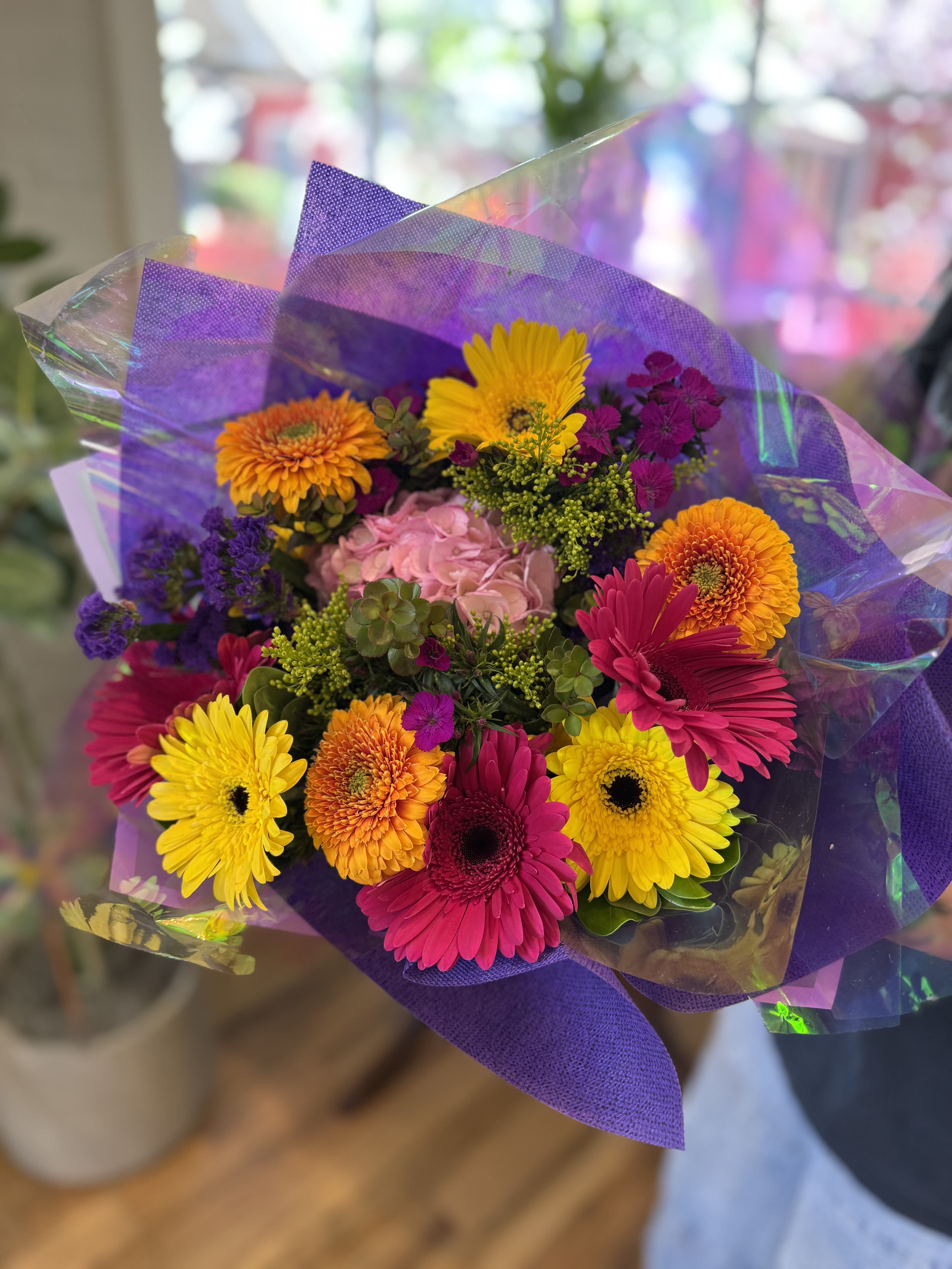 Bright hand-tied bouquet of pink, yellow, and orange gerbera daisies with mixed blooms in purple wrap
