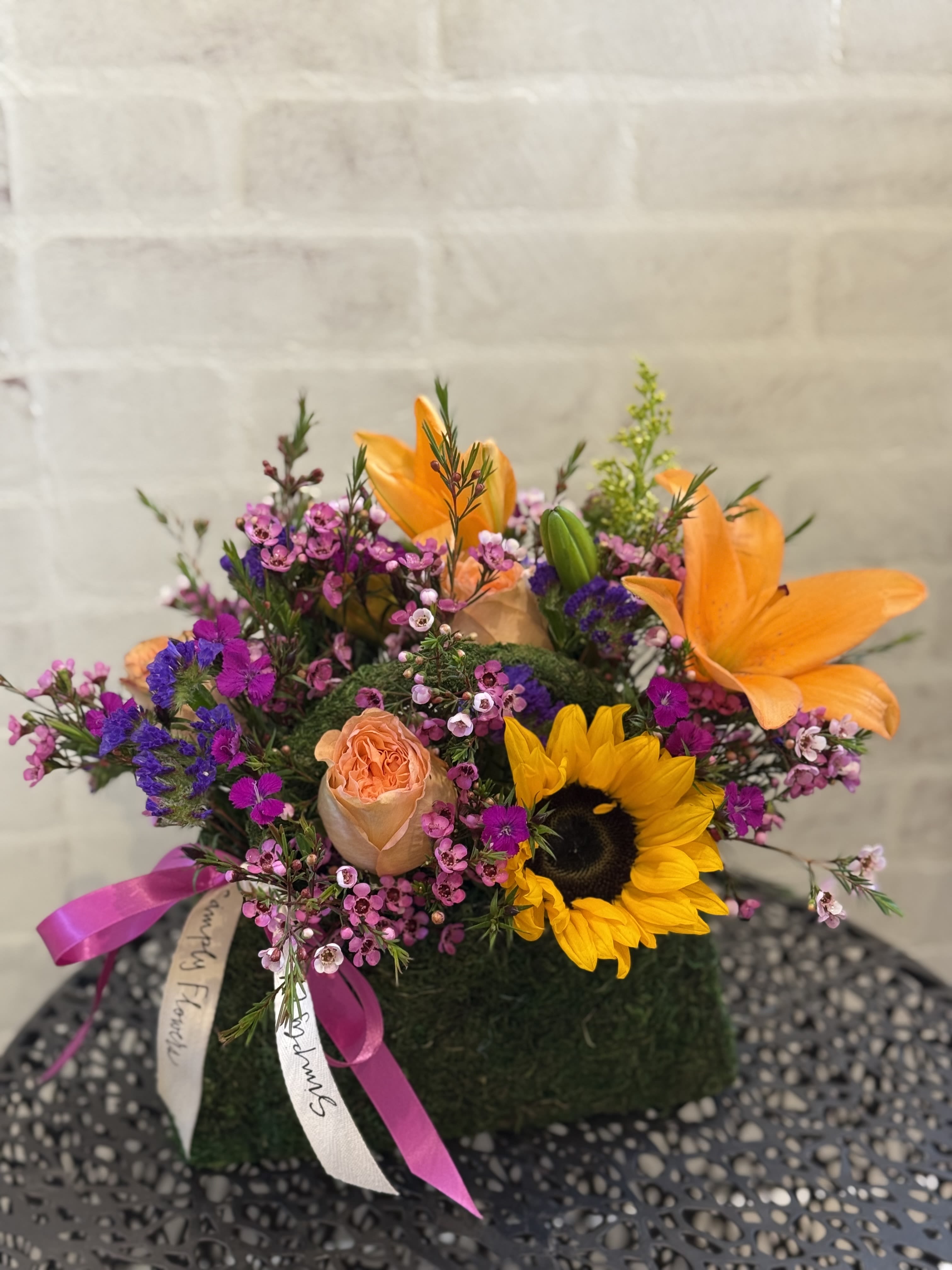 Short mossy cube arrangement with orange lilies, sunflower, peach roses, and pink filler flowers with ribbons.