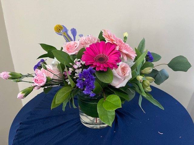 Low glass vase of pink gerbera daisy, roses, and mixed blooms with purple and yellow accents.