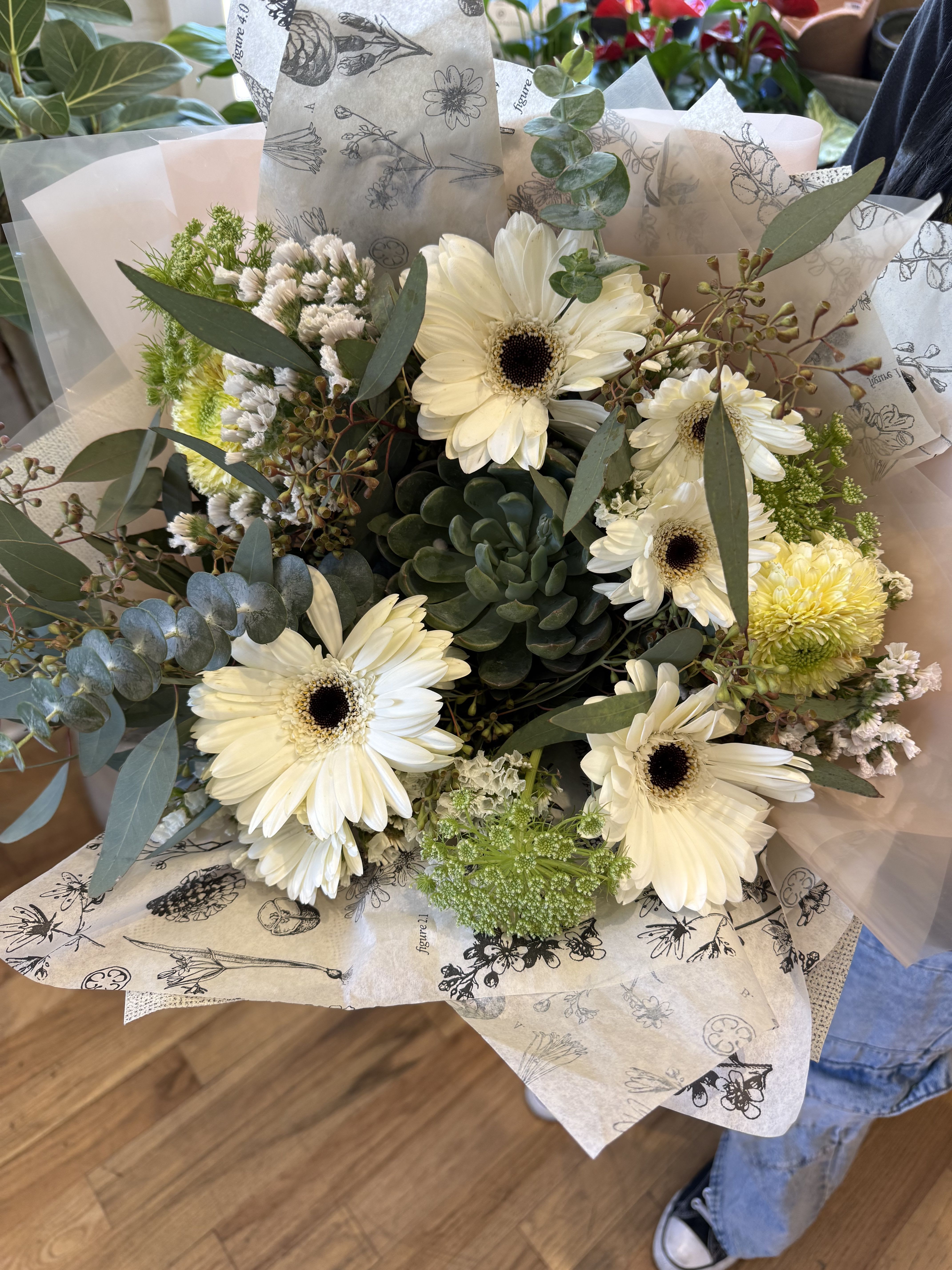 Bouquet of white gerbera daisies with a green succulent center, wrapped in printed tissue and cellophane.