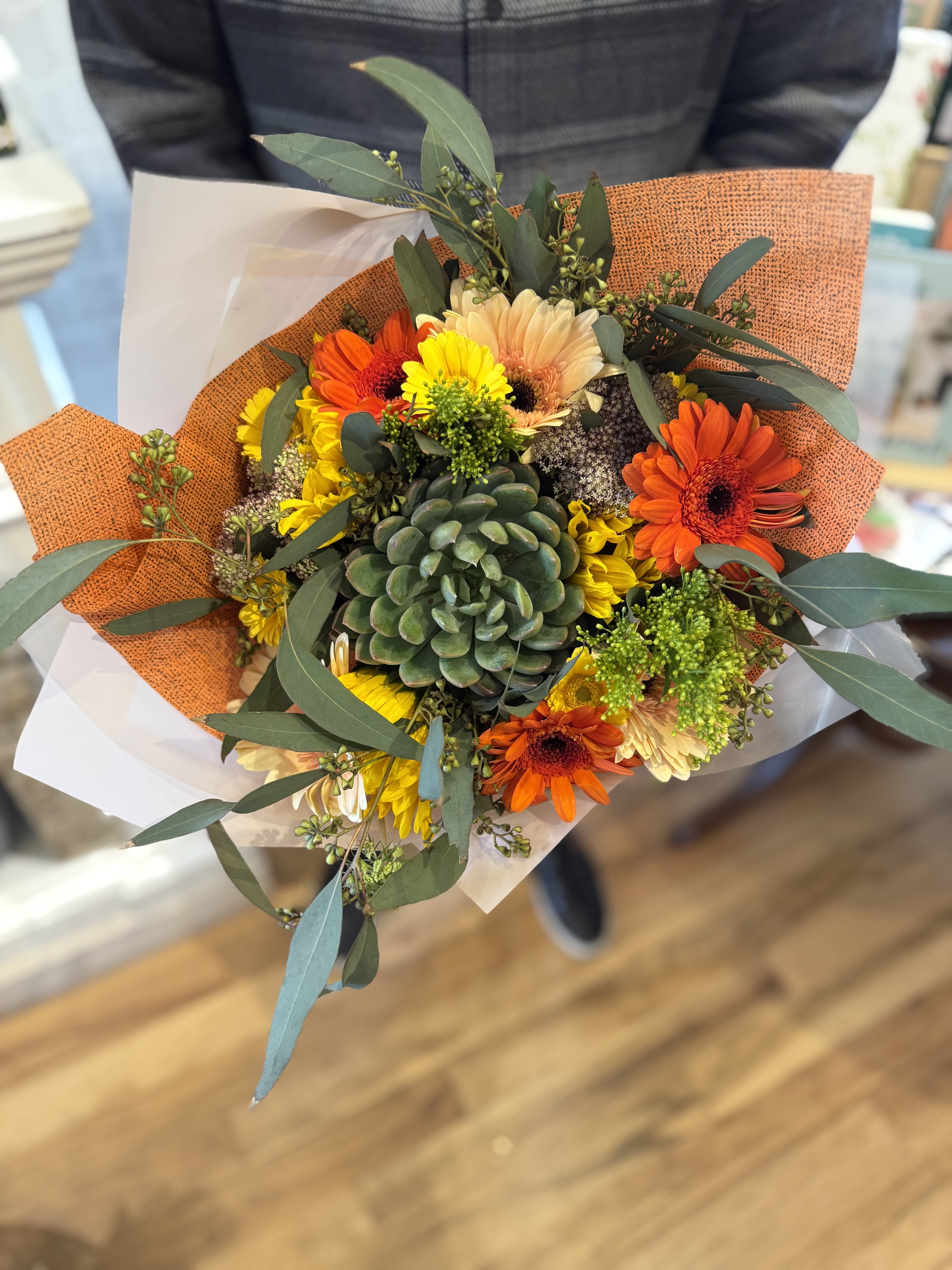Hand-tied bouquet with a central green succulent and orange and yellow gerbera daisies in orange wrap