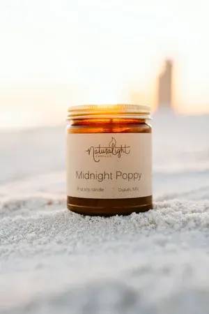 Amber jar candle labeled Midnight Poppy sitting on sand at sunset