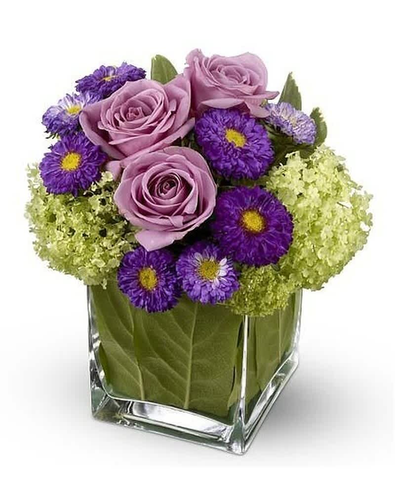 Compact cube arrangement of lavender roses, purple asters, and green hydrangeas in a glass vase lined with leaves.
