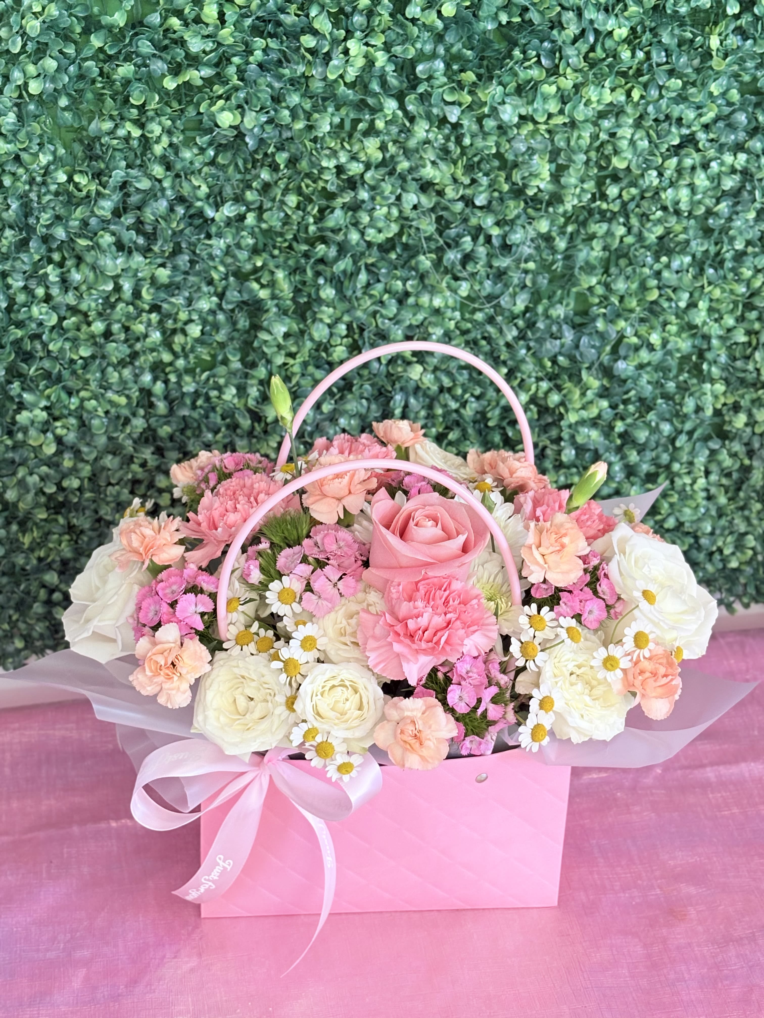 Pink Bloom Bag - Soft pink roses and clusters of creamy white blooms are gathered into a softly rounded floral arrangement-perfect as a birthday bouquet, Mother's Day gift, or a "just because" surprise. The Pink Bloom Bag reads as gentle and celebratory, a feminine flower arrangement that sends a message of care and affection for occasions from graduations and housewarmings to thinking-of-you moments and romantic gestures.  Tightly massed spray roses, carnations, dianthus and tiny white daisies with sunny yellow centers create a full, textured bouquet in a pink gift-bag style container. Pink handles, white tissue wrap peeking at the edges, and a pink satin ribbon bow finish the presentation, giving the design a polished, gift-ready look. Sized to sit low and compact, this pastel arrangement is an easy, elegant choice for saying thank you, sorry, congratulations, or simply brightening someone's day in spring, summer, or any season.