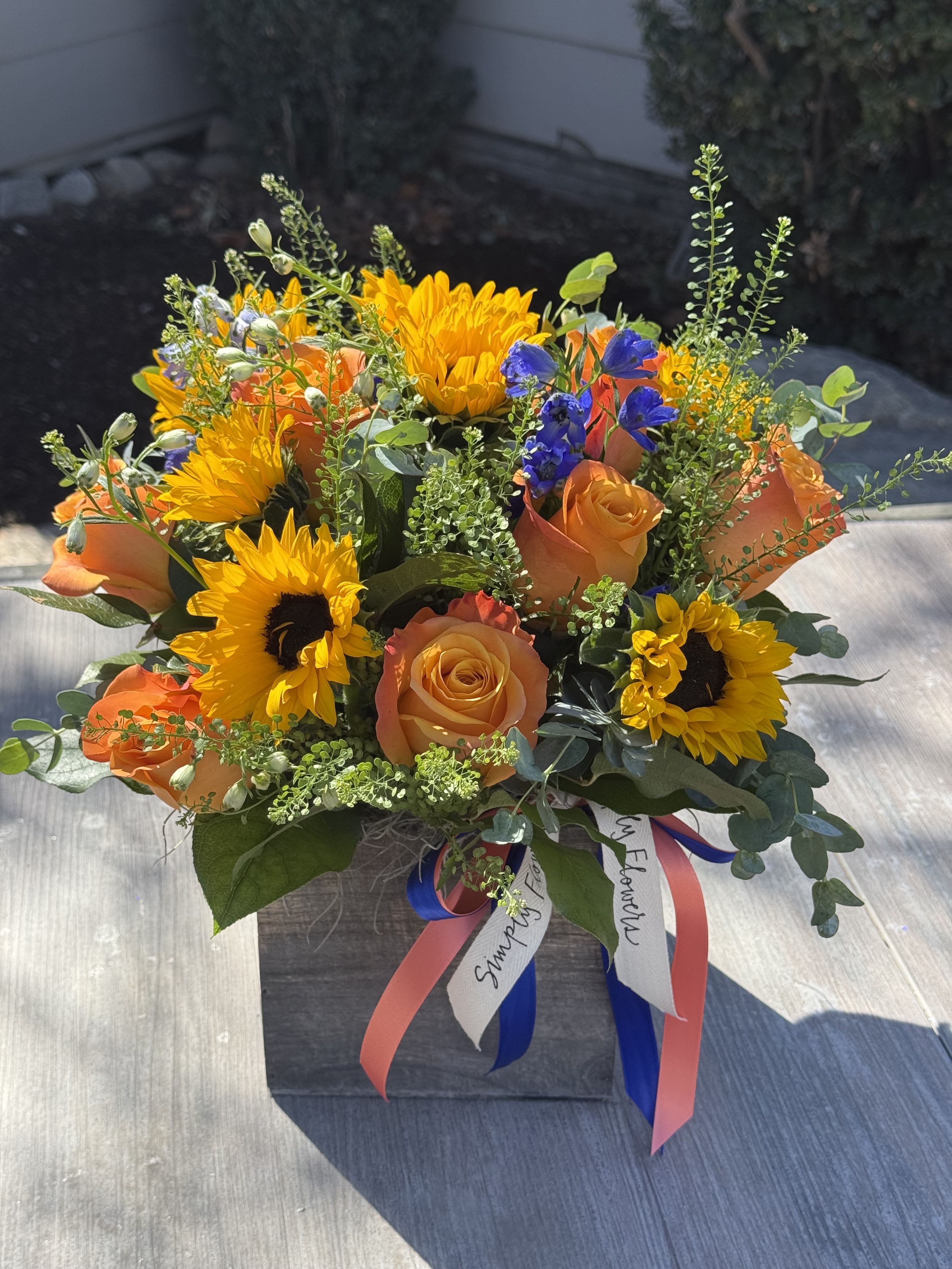 Square wooden box arrangement with yellow sunflowers, orange roses, and blue accent flowers with ribbons