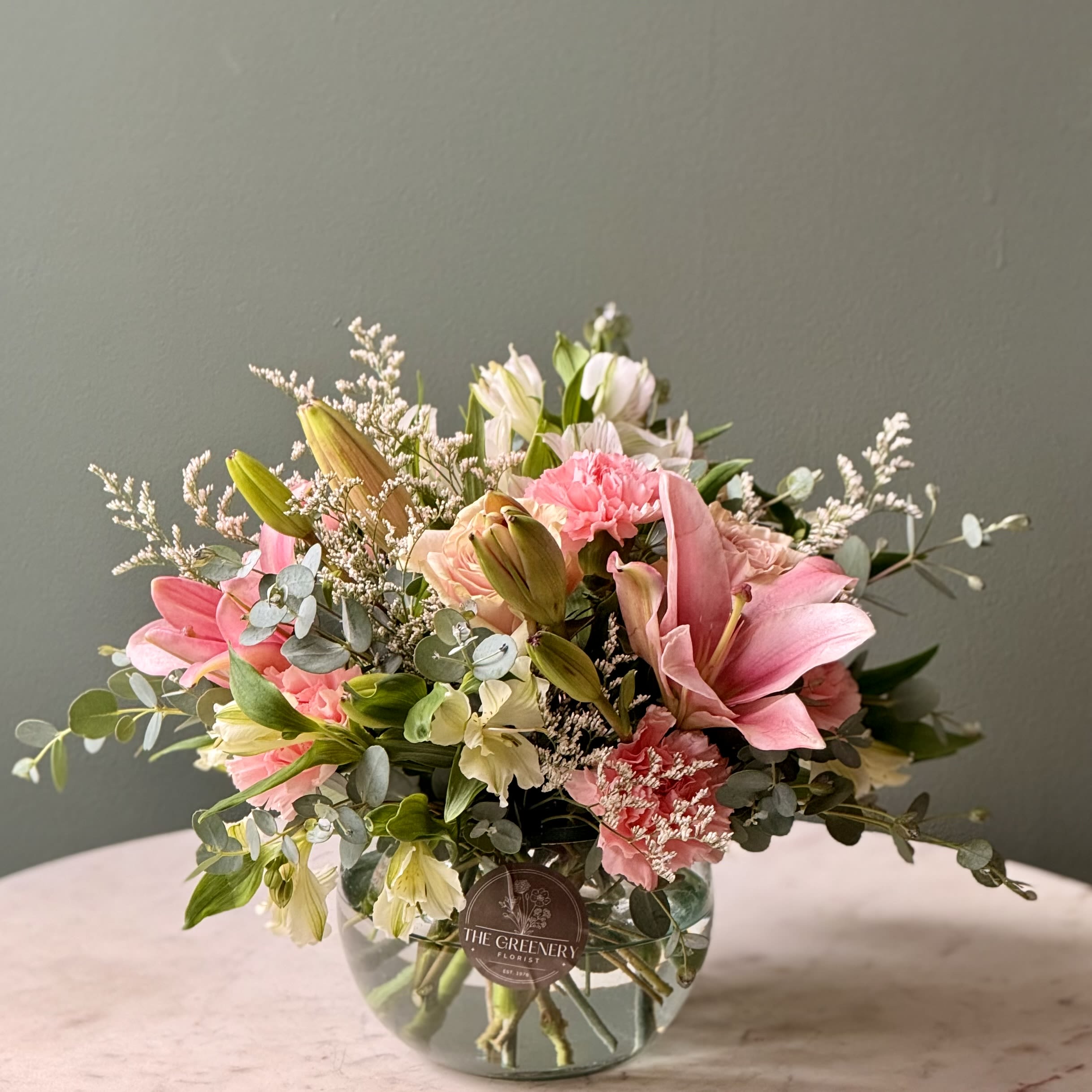 Low glass vase of pink lilies, carnations, and cream alstroemeria arranged in a rounded bouquet.