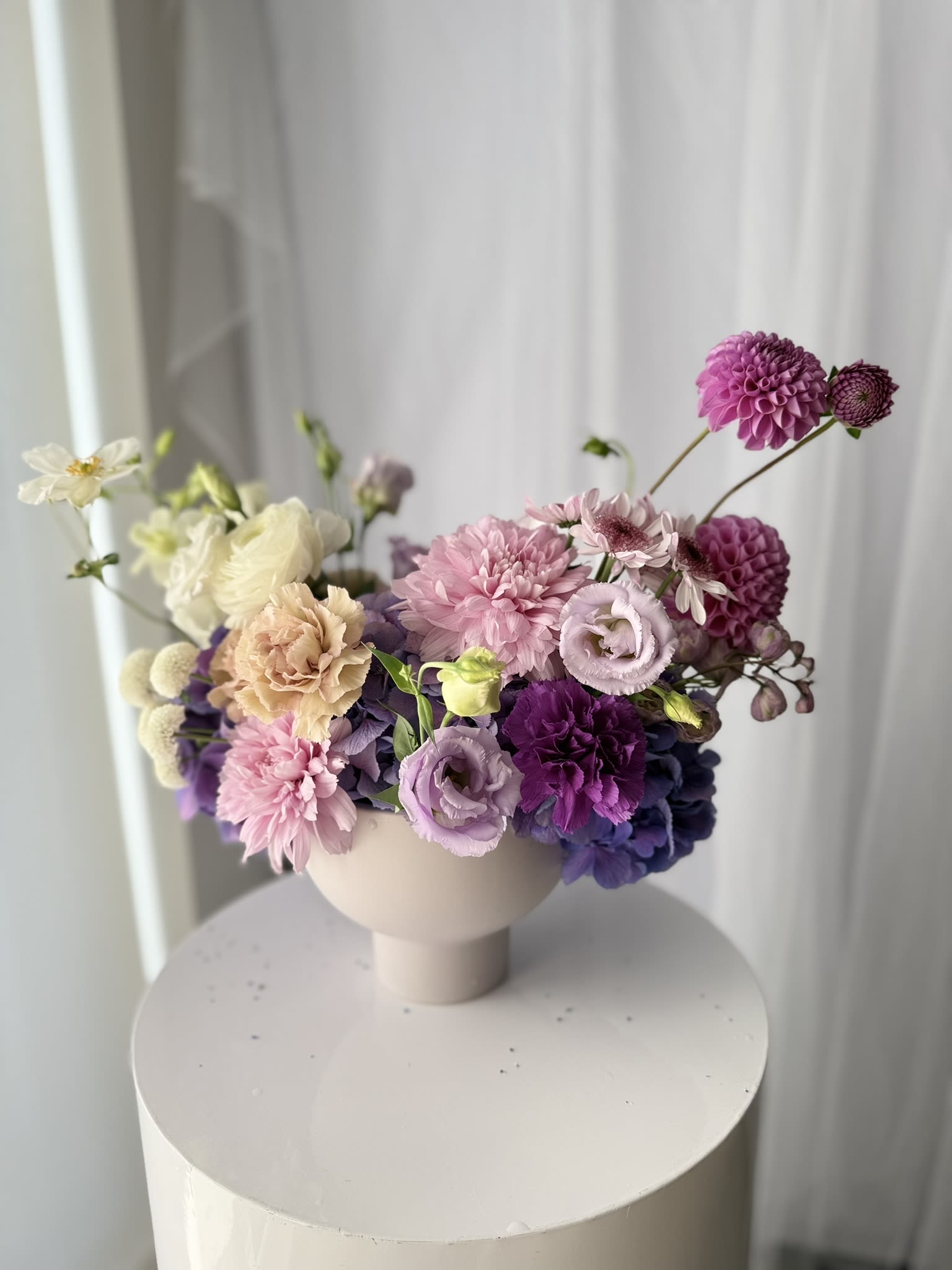 Pastel Garden arrangement - Set in a modern pedestal vase, each stem is placed with intention, creating movement, texture, and a sense of quiet elegance. From the sculptural dahlias to the gentle ruffles of lisianthus and carnations, every detail invites you to pause and appreciate its beauty.  Perfect for intimate celebrations, thoughtful gifting, or bringing a touch of artistry into your everyday space.  Please note: Flower varieties and tones may vary slightly depending on seasonal availability, while maintaining the overall aesthetic and color palette.