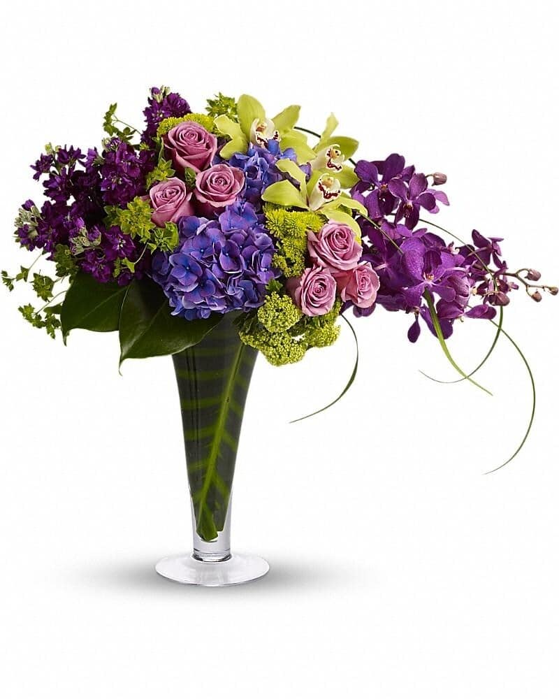 Tall modern arrangement of purple orchids, blue hydrangea, green orchids and lavender roses in a clear vase