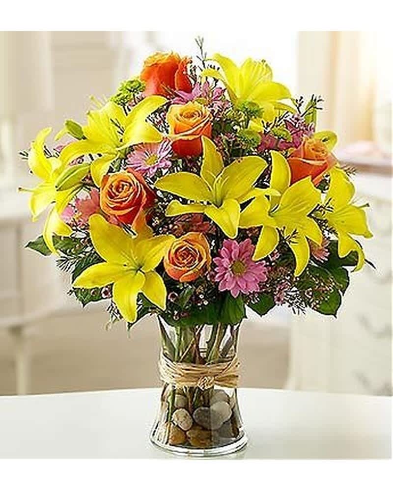 Yellow lilies, orange roses, and pink daisies arranged in a clear glass vase with pebbles