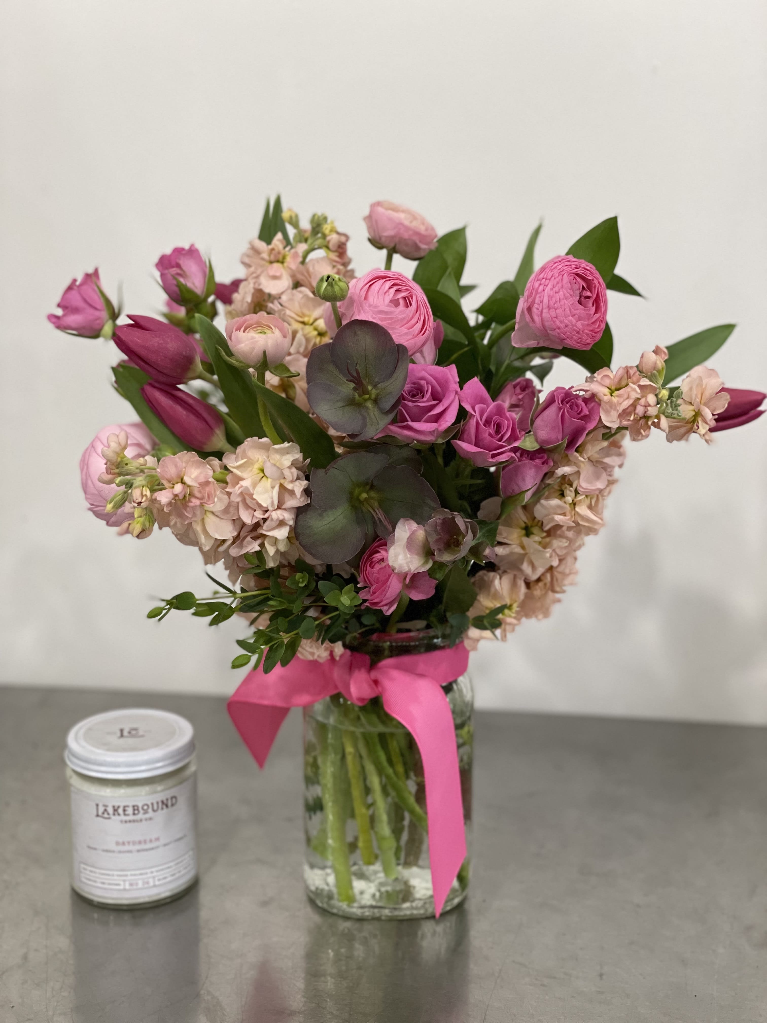 Pink flower arrangement with roses, tulips, and ranunculus in a glass jar with pink ribbon beside a white candle.