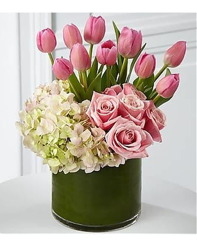 Pink tulips, roses, and pale hydrangeas arranged in a clear glass cylinder vase wrapped with green inside.