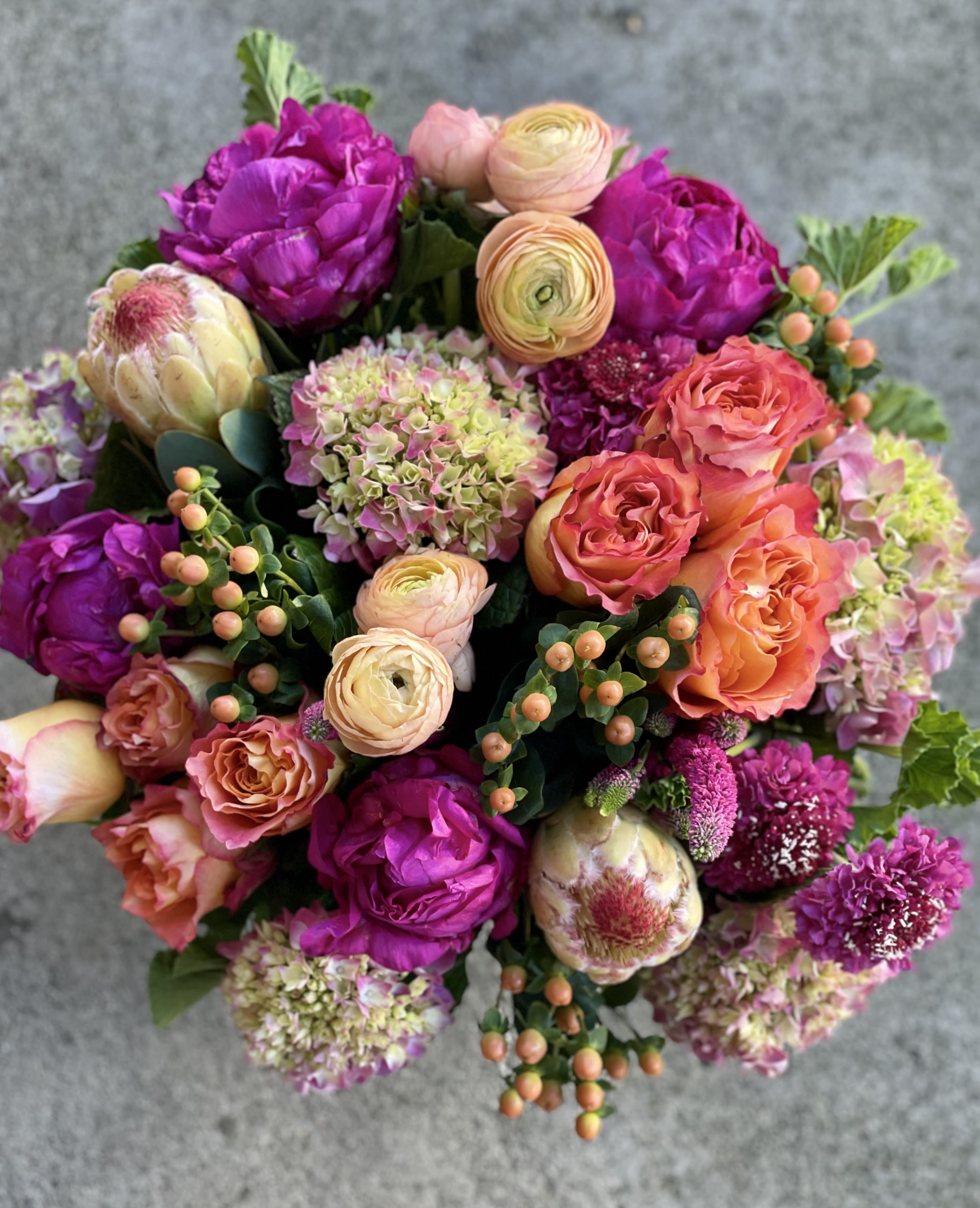 Vibrant round arrangement of fuchsia peonies, orange roses, peach ranunculus, hydrangeas, and protea.