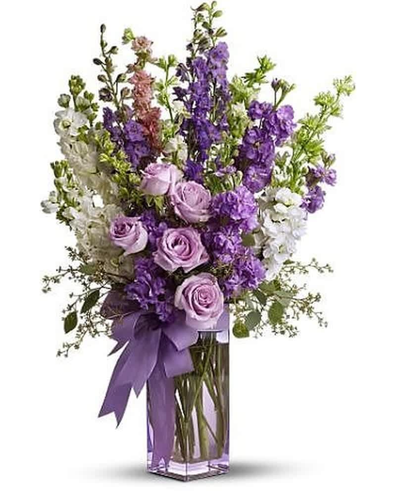 Tall arrangement of lavender roses and purple and white flowers in a clear glass vase with a purple bow