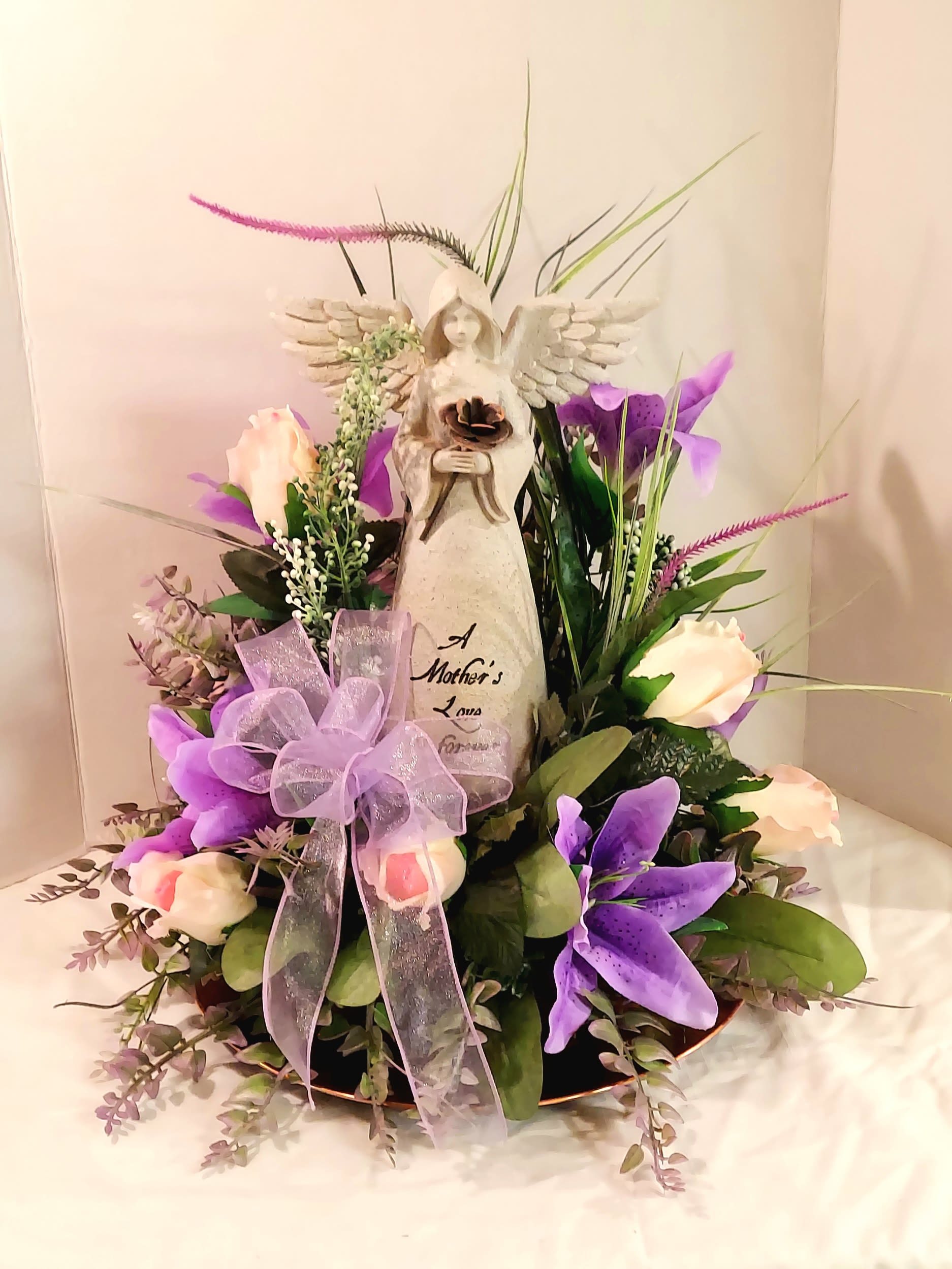 Mother's Love - A pale stone angel takes center stage in this touching flower arrangement, making it an especially meaningful choice for a Mother's Day bouquet, sympathy tribute, or a comforting keepsake to honor a mother. The circular design of the arrangement and the soft palette of purple, white, pink, and green create a gentle, reflective impression that will help the person you're thinking of feel tenderly remembered and cared for.  Cream roses with blush centers and soft purple lilies are arranged around the angel in a low, round container, accented by wispy spikes and textured green foliage for subtle movement and depth. A sheer lavender ribbon bow at the front echoes the purple tones and adds an airy, delicate finish. The angel figurine holds a small flower and bears a sentiment about a mother's love, making this piece both a floral gift and a lasting memento.  Ideal for a mother, grandmother, or someone you want to comfort, this arrangement reads as heartfelt and personal whether given for a birthday, anniversary, memorial, or simply to say you're thinking of them. Sized to sit gracefully on a table or mantel, it arrives ready to display-just add fresh water and enjoy the gentle fragrance of lilies and roses as the keepsake offers quiet reassurance.
