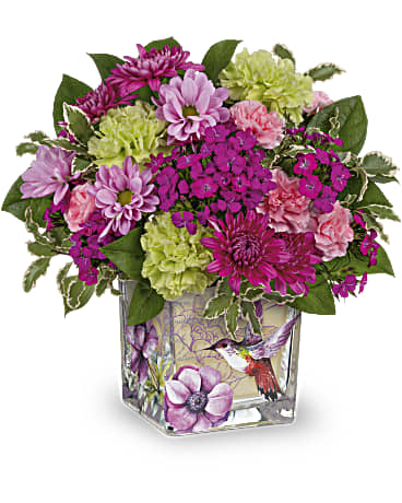 Compact arrangement of purple mums and green and pink carnations in a square hummingbird vase