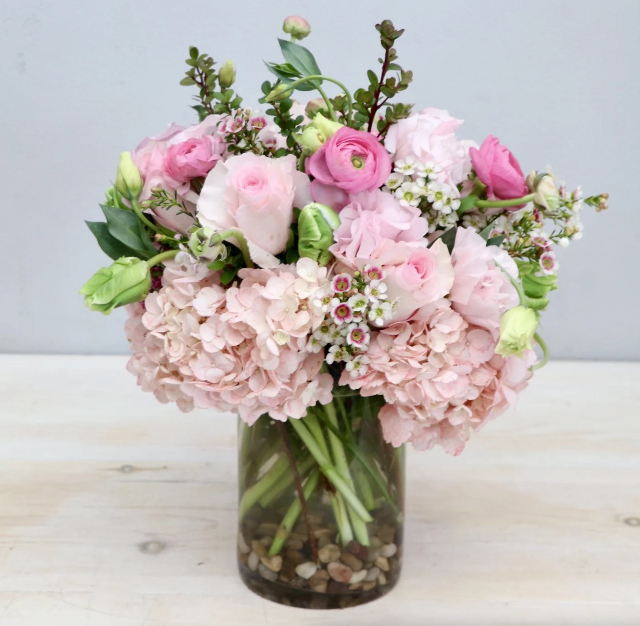 Pastel pink hydrangeas, roses, and ranunculus arranged in a clear glass vase with pebbles at the base