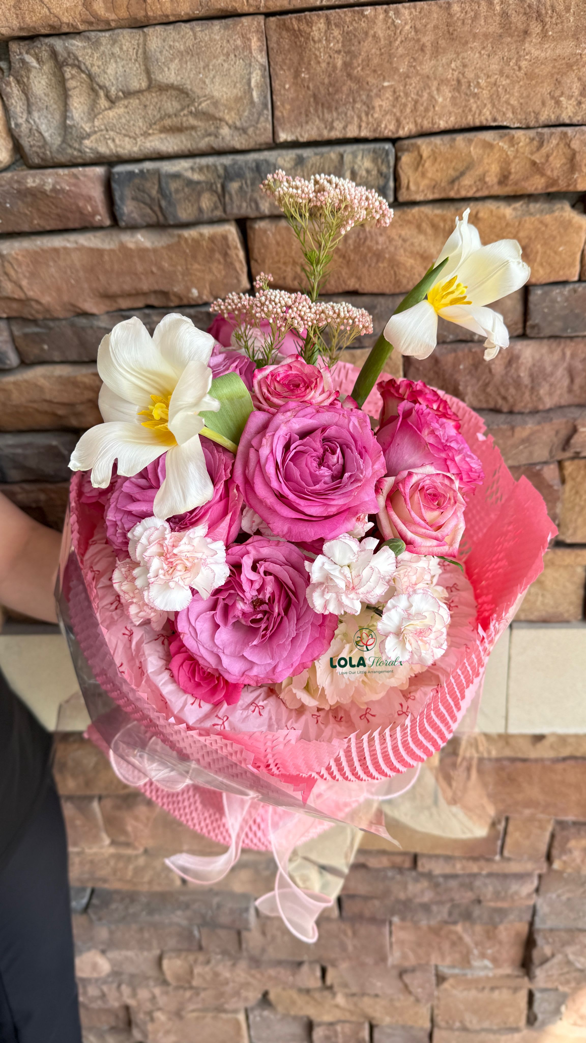 Lovely Pink Rose Bouquet  - A dense cluster of hot pink roses makes this hand-tied bouquet a joyful choice for a birthday bouquet or an anniversary roses gift that feels both romantic and playful. The vivid pink palette-anchored by bold roses and softened by blush-pink carnations-creates an immediate, celebratory impression the person you're thinking of will notice the moment it arrives.  Two open white tulips with sunny yellow centers rise above the pink mass to add bright contrast, while airy sprays of small pink filler blooms lend delicate texture. Wrapped in layered pink floral paper, pink mesh, and clear cellophane, and finished with a soft sheer ribbon bow, the arrangement reads as thoughtfully finished and easy to carry. Sized as a rounded, hand-tied bouquet, it's a meaningful way to say congratulations, happy birthday, or simply that you care-no special care required beyond fresh water and a clean vase to keep the flowers looking their best.