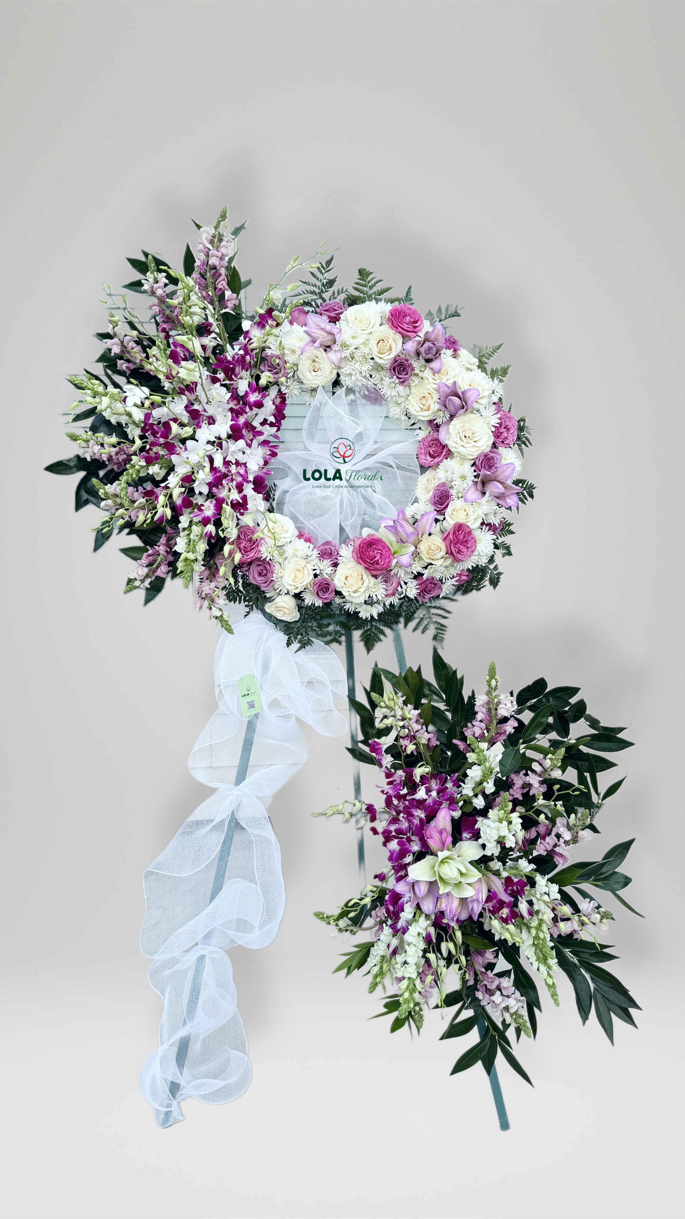 Eternal Peace - A softly rounded wreath of white and pink roses with white chrysanthemums and pale lavender blooms sets a serene, contemplative tone-perfect for a standing sympathy arrangement for a funeral or a gentle way to say "thinking of you" after a loss. The design's sweeping cluster of purple and white dendrobium orchids and snapdragons adds vertical movement and quiet elegance, making it an appropriate memorial display for Memorial Day services or any remembrance gathering.  This standing arrangement is mounted on a metal easel with a wreath frame and finished with a white mesh ribbon bow and a printed center sign for a respectful, formal presentation. A coordinating spray of orchids, snapdragons, and lilies is placed beneath the wreath on the same easel to visually ground the piece; colors range from soft white and pink to lavender and deeper purple accents for depth and contrast. The mix of roses, chrysanthemums, dendrobium orchids, lilies, and snapdragons creates varied textures-from the soft rose petals to the sculptural orchids-so the recipient's memory is honored with thoughtful, enduring beauty.  Designed to convey sympathy and calm, this arrangement is the kind of tribute that makes mourners feel seen and comforted. It arrives ready to display on its easel at a service or graveside; if you need sizing or delivery specifics for a particular venue, please let us know so we can ensure it fits the setting.