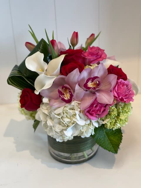 Compact arrangement of white hydrangeas, red roses, pink orchids, tulips, and calla lilies in a glass vase.