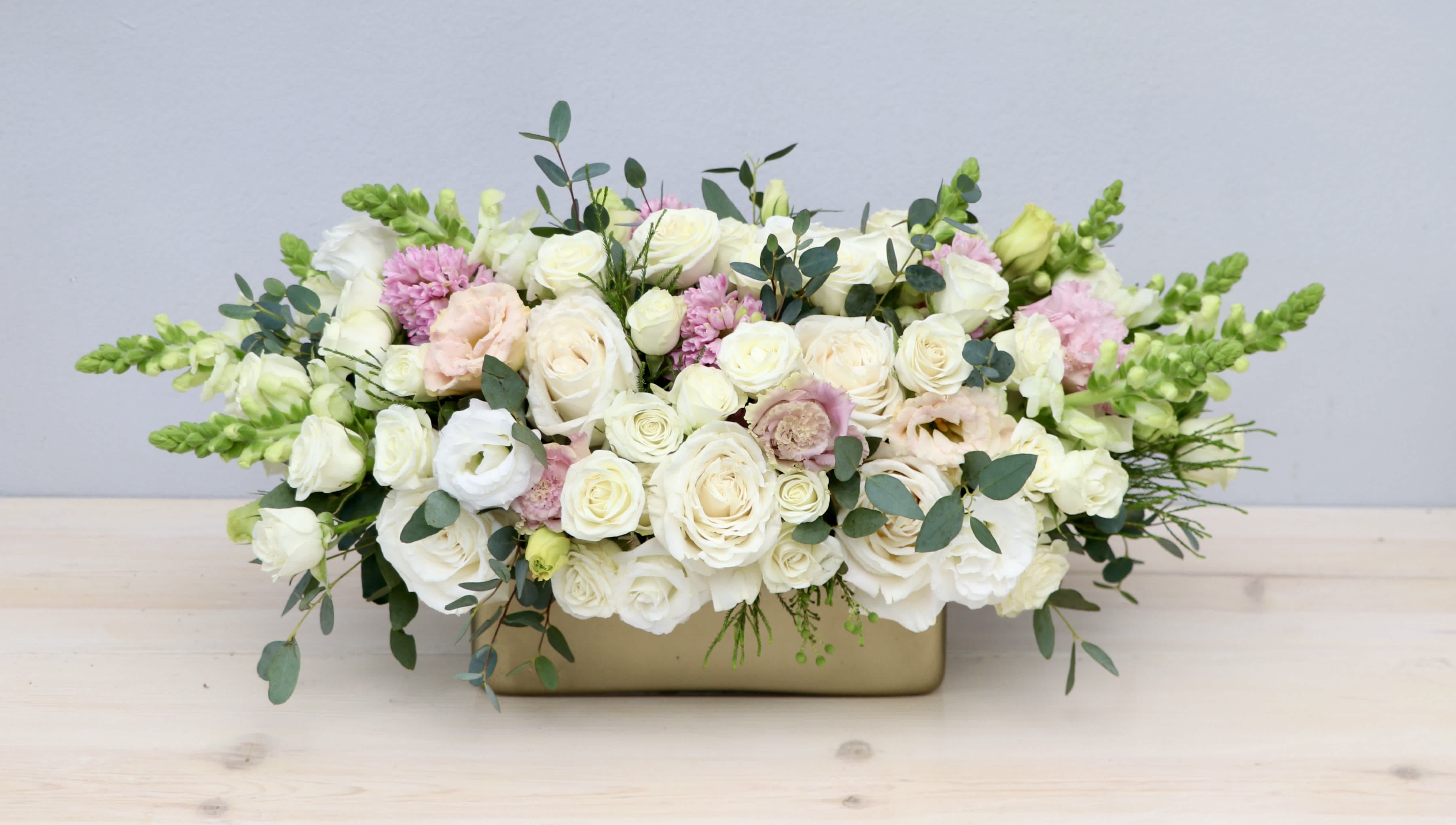 Low centerpiece of white and blush roses with green snapdragons in a gold rectangular vase