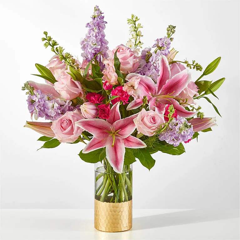 Pink lilies, roses, and lavender stock arranged in a clear glass vase with a gold base