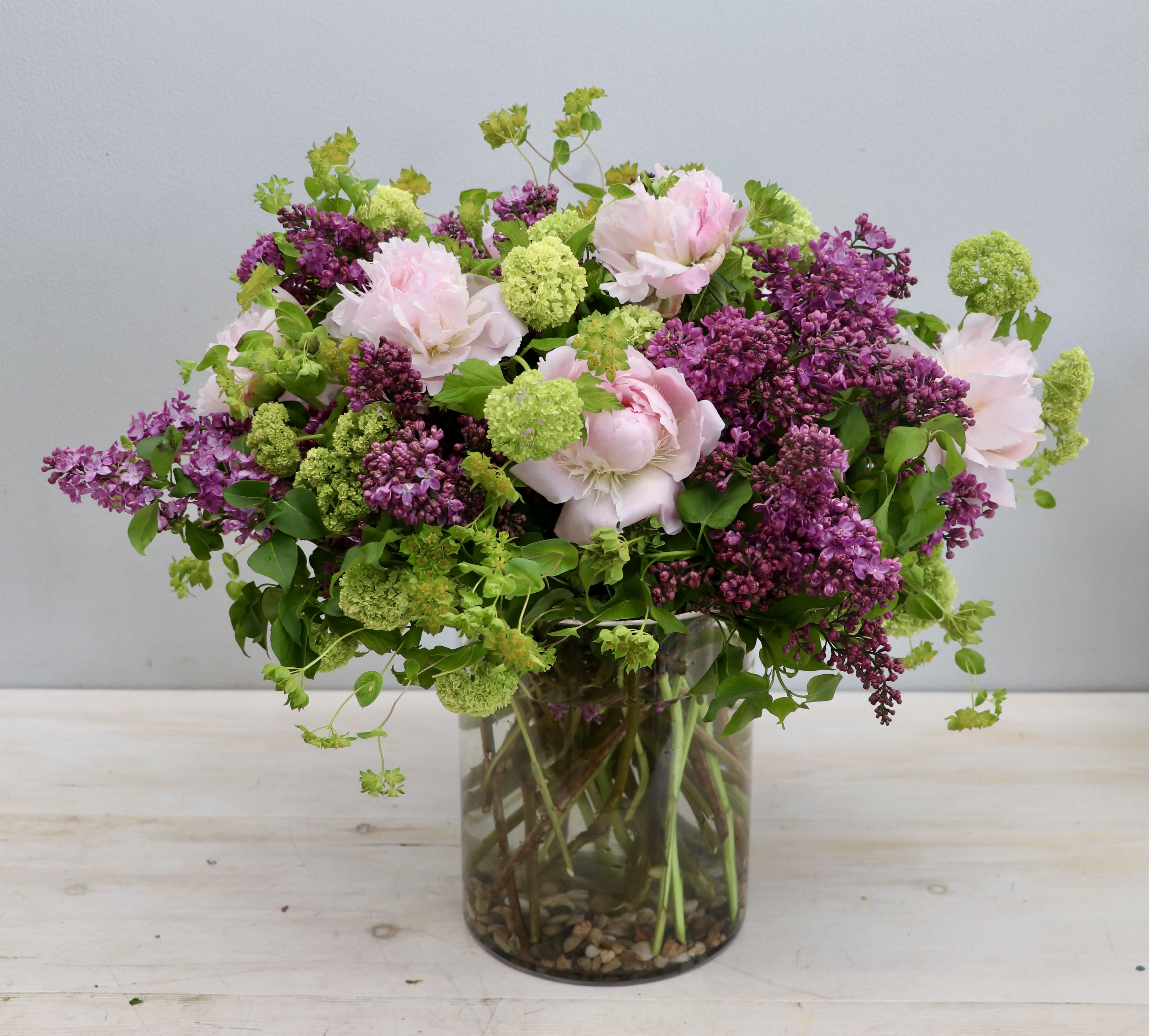 Loose garden-style bouquet of pink peonies, purple lilacs, and green blooms in a clear glass cylinder vase