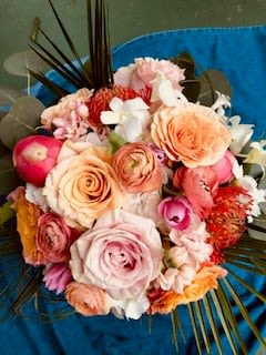 Just Peachy - A compact, round flower arrangement that greets the eye with warm peach and blush roses paired with delicate ranunculus-style blooms - perfect as a Mother's Day bouquet or for anniversaries, housewarmings, and just-because surprises. Bright orange pincushion protea and vivid pink accents lift the palette, while a few white blossoms keep the look fresh and light. The overall effect is cheerful and elegant, the kind of gift that makes the person you're thinking of feel genuinely celebrated.  Textured palm fronds and other foliage fan from the back to give the design a slightly tropical, modern character that reads as both relaxed and refined. Colors include peach, pink, yellow, white, and orange; flowers feature roses, ranunculus-type blooms, protea, and complementary seasonal stems. Ideal for occasions from get well and thank you to sympathy, retirement, weddings, and expressions of love or apology - a versatile arrangement that looks thoughtfully composed on a bedside table, entry console, or dining centerpiece.