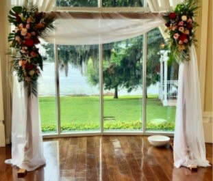 Tropical Vibe Wedding Arch - A wooden wedding arch wrapped in soft white sheer fabric that pools gently on the floor creates an immediate sense of airy romance - the perfect floral backdrop for a wedding ceremony or vow renewal. Dense, asymmetrical clusters of red, pink, peach and white roses and spray roses crown the upper left and right corners, so the person you're celebrating will feel seen and cherished the moment they step beneath it. The contrast of vivid blooms against flowing white drape gives a slightly formal, timeless look.  This design pairs classic roses and spray roses with abundant textured greenery, arranged to extend outward for a garden-inspired silhouette. Sized to serve as a ceremony focal point, it suits indoor or outdoor spaces with tall windows or garden views; it's ideal for weddings, anniversaries, and vow renewals when you want the recipient to feel celebrated, romantic, and warmly embraced.