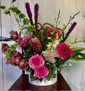 Pretty in Pink - Hot pink roses and gerbera daisies take center stage in this Pretty in Pink flower arrangement, a birthday-ready and Mother's Day-appropriate bouquet that feels both celebratory and thoughtful. The low, clustered pink blooms sit in a clean white ceramic cylinder vase while taller purple spike flowers and Bells of Ireland lift the design into a garden-like silhouette-perfect for saying "happy birthday," "congratulations," or simply "just because" to someone you care about.  At the heart, a textured tropical bloom and arching pale dendrobium orchids introduce an exotic contrast in shape and color, while fresh green accents and white highlights keep the palette lively and balanced. The mix of roses, gerbera daisies, snapdragons, orchids and Bells of Ireland gives the arrangement varied height and texture, making it a bright centerpiece for a bedside table, office desk, or kitchen counter.  Send this arrangement as a cheerful get-well gift or a warm thank-you-she'll feel genuinely celebrated by the bold pinks and uplifting verticals. Presented in a sturdy white ceramic vase, it arrives ready to display; refresh the water and trim stems every few days to keep blooms at their best.
