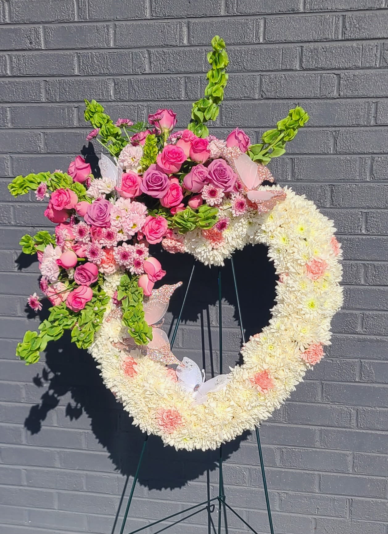 Standing Heart  - A tender heart-shaped standing spray in soft pinks, whites, lavender accents and fresh greens, designed as a sympathy heart arrangement to say "you are remembered" with quiet grace. This standing heart uses plush white chrysanthemums and pale pink carnations to create a cushiony, comforting base, while clusters of pink roses and small pink-and-white daisies gather on the upper left for a focal lift-the result feels intimate and thoughtfully arranged for funerals, memorials, or a service honoring a loved one.  The composition also includes tall Bells of Ireland that arch outward for gentle height and movement, plus lilies and red roses woven into the design for subtle contrast and depth. Decorative butterfly picks tucked among the blooms add a delicate, hopeful touch. The tribute is presented on a sturdy metal easel (wire easel option available), giving it a formal display presence suitable for chapels or graveside services.  Ideal for a close friend, family member, or someone who brought light to your life, this standing heart communicates love, remembrance, and gentle sympathy. It arrives ready to place on the easel; to keep the flowers looking their best during services, avoid direct sun and check water source if provided by the venue.