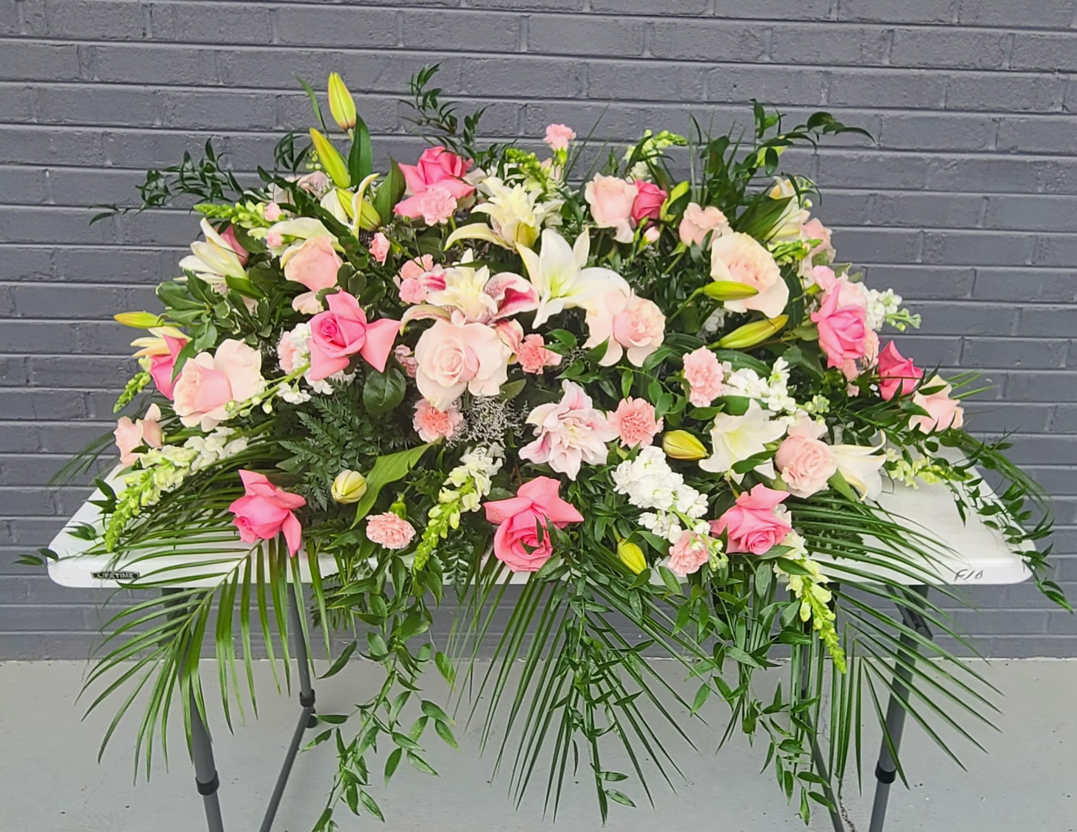 Casket Spray - Soft blush roses and creamy white lilies form a tender, elongated casket spray designed for sympathy and funeral settings, offering a gentle, comforting presence for the person you're honoring. Pink carnations and clusters of pale roses create a warm, affectionate center while white snapdragons and stock add upright texture through the middle-together they read as a calm, reassuring tribute that will help the recipient feel remembered and cared for.  This wide, low arrangement measures as a large, cascading spray with open and budded lilies rising just above dense rose and carnation clusters. Long palm fronds and trailing greenery extend from the ends for a sweeping profile suited to display on a service table or white folding table, and the pastel pink, white, green, and soft yellow accents create a refined, feminine palette appropriate for memorials, moments honoring mothers, or other gentle commemorations.