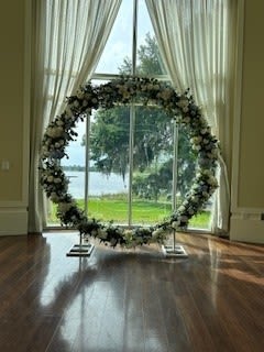 Round Arch with full flower coverage - A grand circular wedding arch wrapped in lush white blooms and deep green foliage creates an unforgettable ceremony focal point - perfect as a wedding ceremony backdrop or romantic photo setting. This round arch with full flower coverage frames the couple with continuous roses and hydrangeas, giving a soft, textured garland that reads both timeless and modern for your special day.  Crafted on a sturdy metal circular arch frame and supported by simple metal stands, the design keeps attention on the floral ring while providing reliable stability indoors or at sheltered outdoor venues. The palette of crisp whites and rich greens complements classic, minimalist, or lakeside weddings and pairs beautifully with sheer draping or natural light for an airy, serene look.  Made to be the visual centerpiece that makes your person feel truly celebrated, this arch suits ceremony aisles, altar spaces, or a dedicated photo moment. Ask about installation, delivery, and sizing options to ensure the arch fits your venue and timeline; care during setup is minimal since blooms are secured to the metal frame for a full, finished presentation.