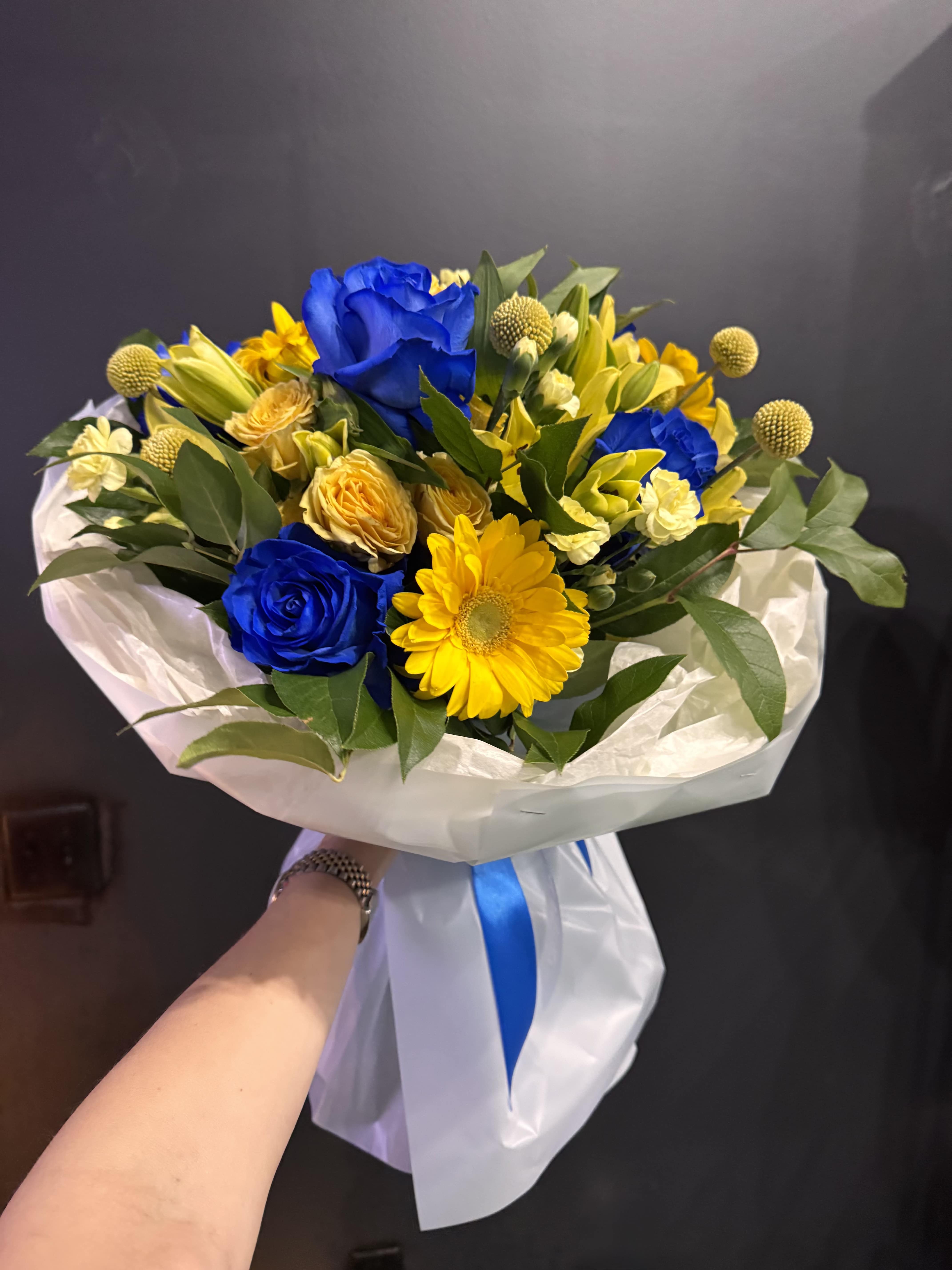 Golden Blue Harmony - Golden Blue Harmony is a striking bouquet that captures attention with its vivid contrast of deep blue roses and radiant yellow blooms. Featuring cheerful gerberas, delicate spray roses, seasonal accents, and lush greenery, this arrangement is designed to uplift and impress. The combination of colors creates a fresh, modern, and joyful look — perfect for birthdays, celebrations, or simply brightening someone’s day. Wrapped in elegant white paper with a soft ribbon, it feels both refined and expressive.  