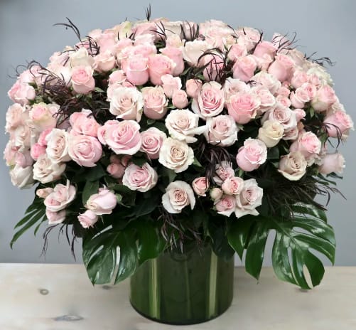 Large dome of pale pink and ivory roses in a green glass cylinder vase with glossy tropical leaves.