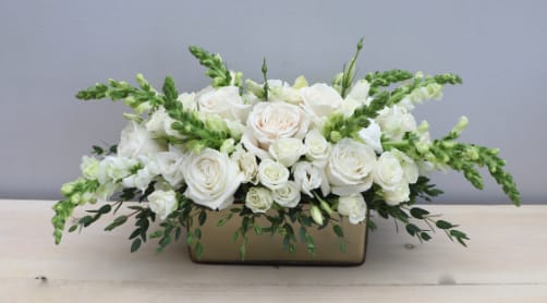 Low rectangular arrangement of white roses and snapdragons in a gold container